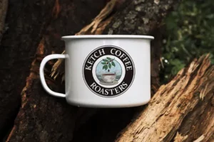 The Ketchikan Coffee Roasters logo printed on the side of a small white coffee cup, lying on its side on some logs and foliage.