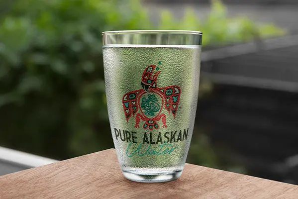 The pure Alaskan Water Logo on a clear glass covered in condensation, sitting on a brown table outside.
