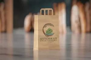The Ketchikan Ever Greens logo is printed on the side of a brown paper bag, sitting on a reflective cocreate countertop.