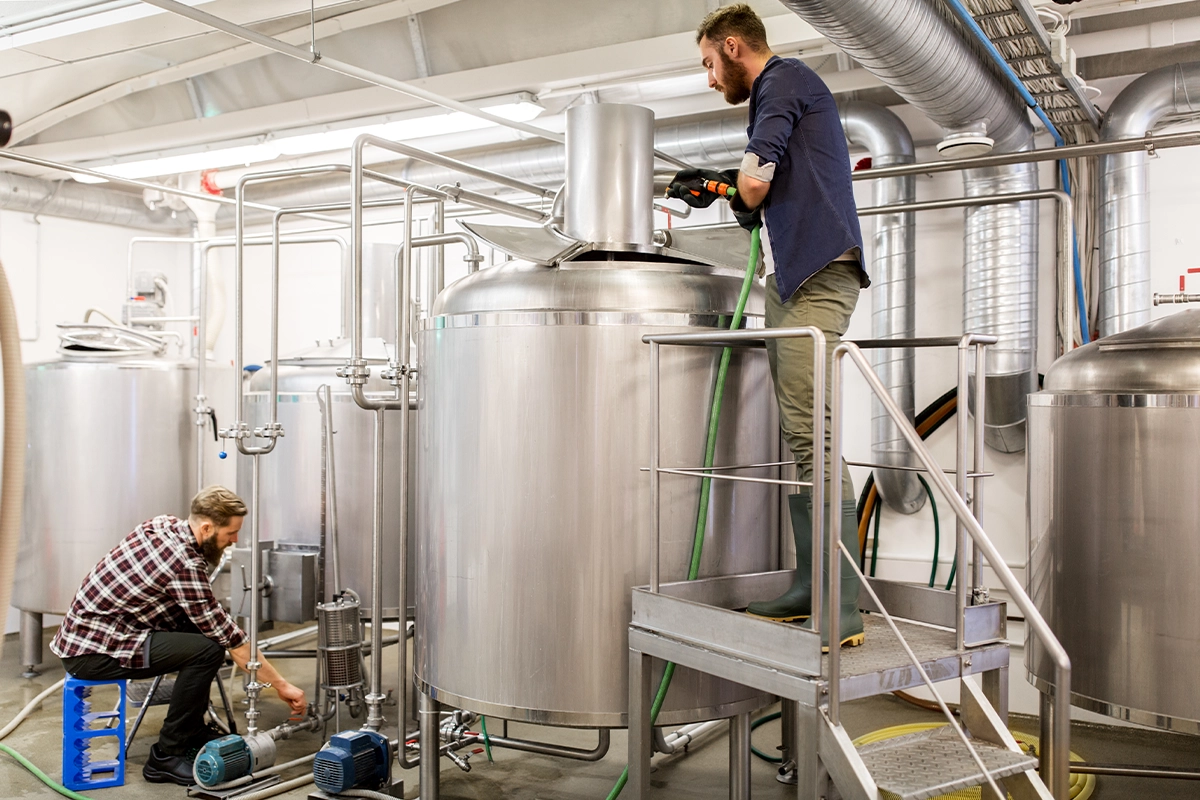 Man Working - Bold Marketing & Design Two workers in a brewery operating large stainless steel fermentation tanks and industrial equipment.