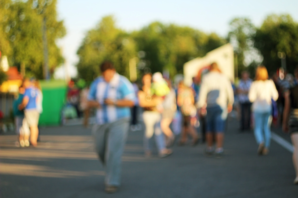 Out-of-focus crowd walking outdoors at a community festival on a sunny day with trees in the background.