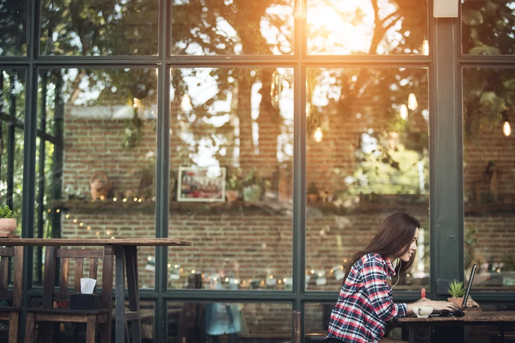 Young woman wearing headphones working on a laptop in a cozy café with warm sunlight reflecting off the window.