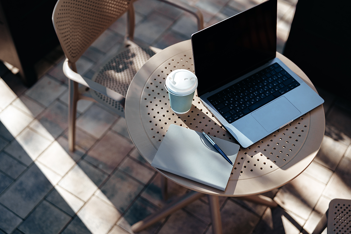 Laptop, coffee cup, and notebook arranged on a small outdoor café table, representing remote work and creative productivity.