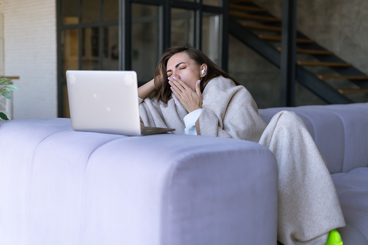Woman wrapped in a blanket yawning while using a laptop on a couch, representing fatigue from poor website experience or digital overload.