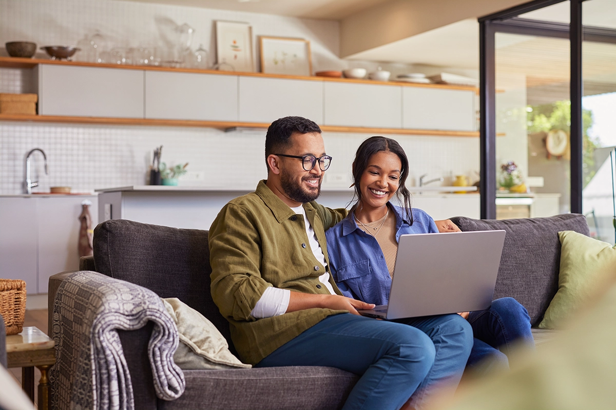 Smiling couple sitting on a couch at home using a laptop to search online together.