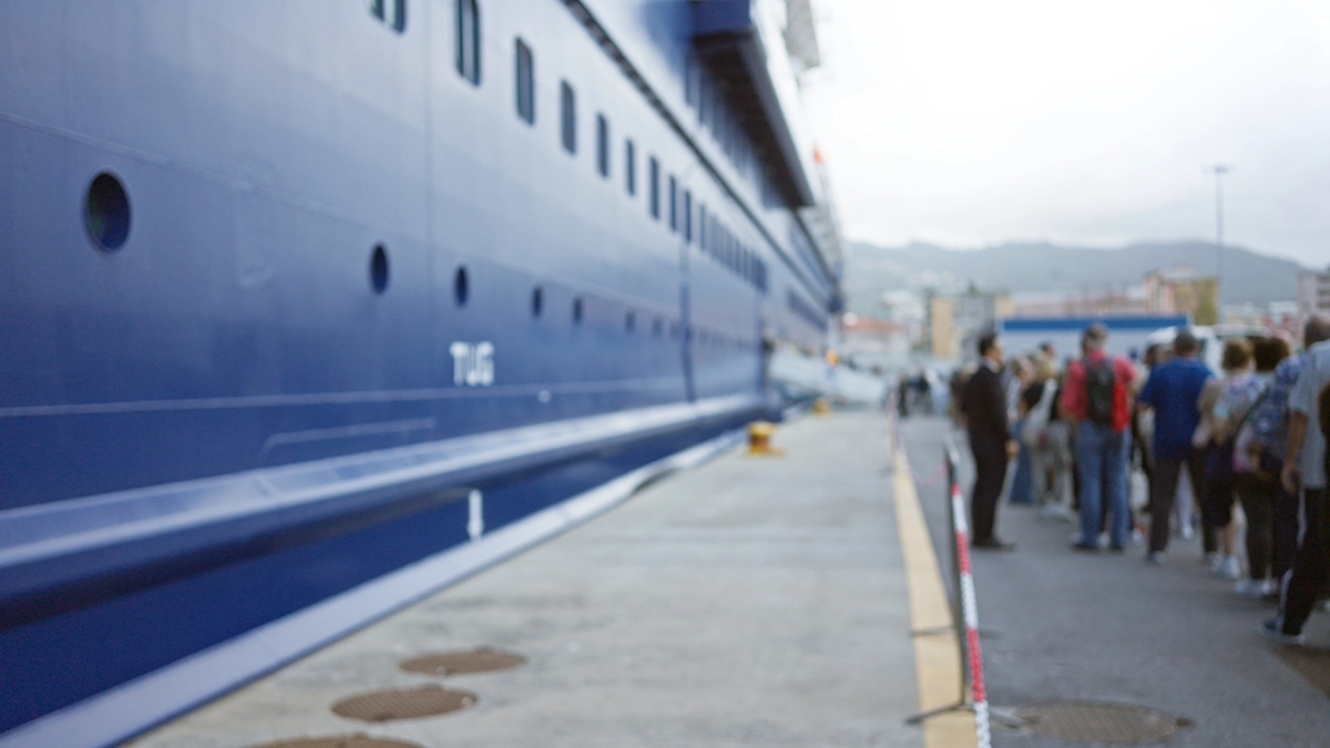 Cruise passengers lining up to board a large blue cruise ship at port on an overcast day.