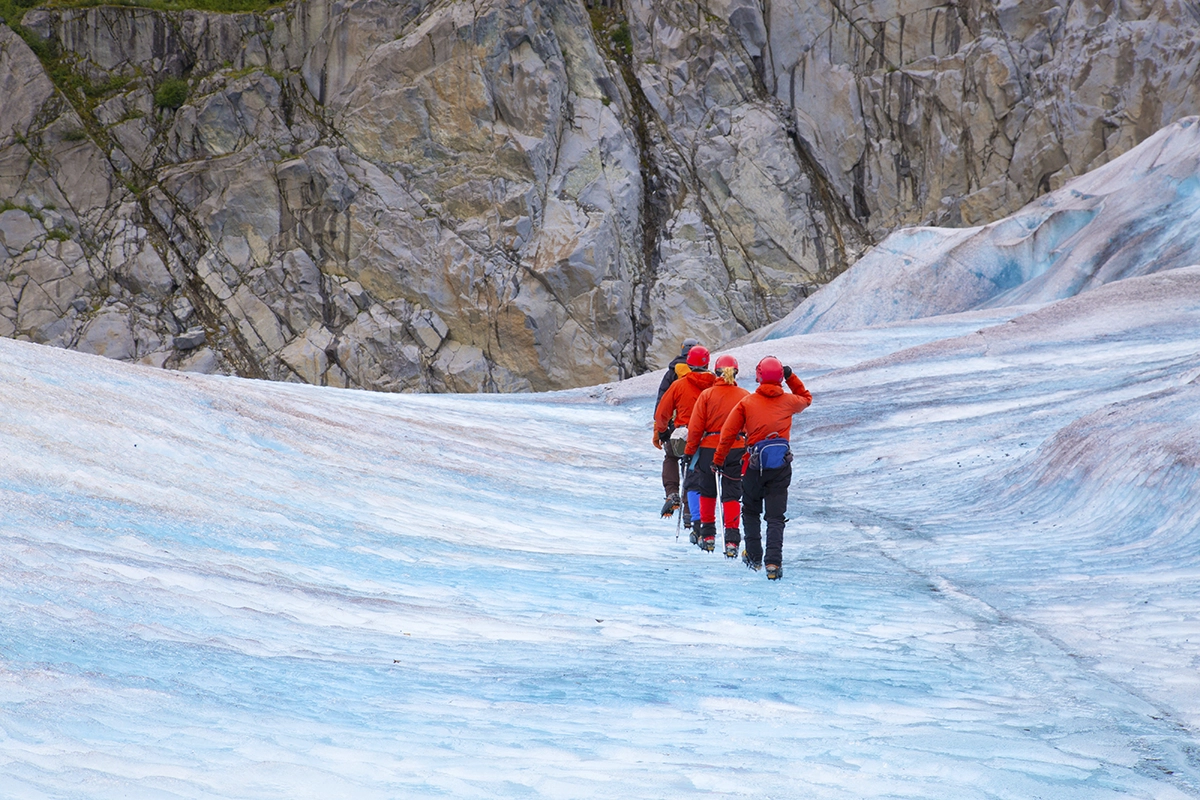 Four hikers in red jackets trekking across Mendenhall Glacier surrounded by rocky cliffs in Alaska.