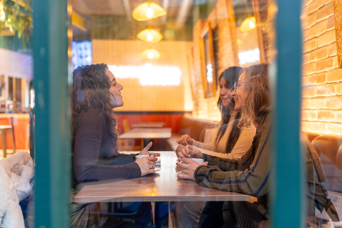 Three women enjoying coffee and conversation inside a cozy modern cafe with warm lighting.