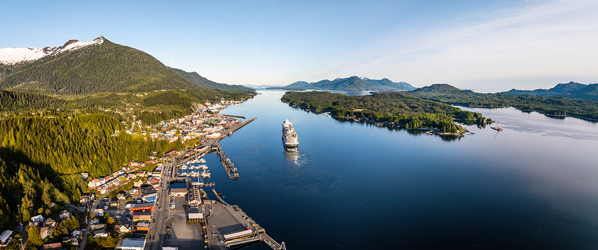 Aerial view of a coastal town surrounded by forested mountains and water, with a cruise ship sailing through the harbor.