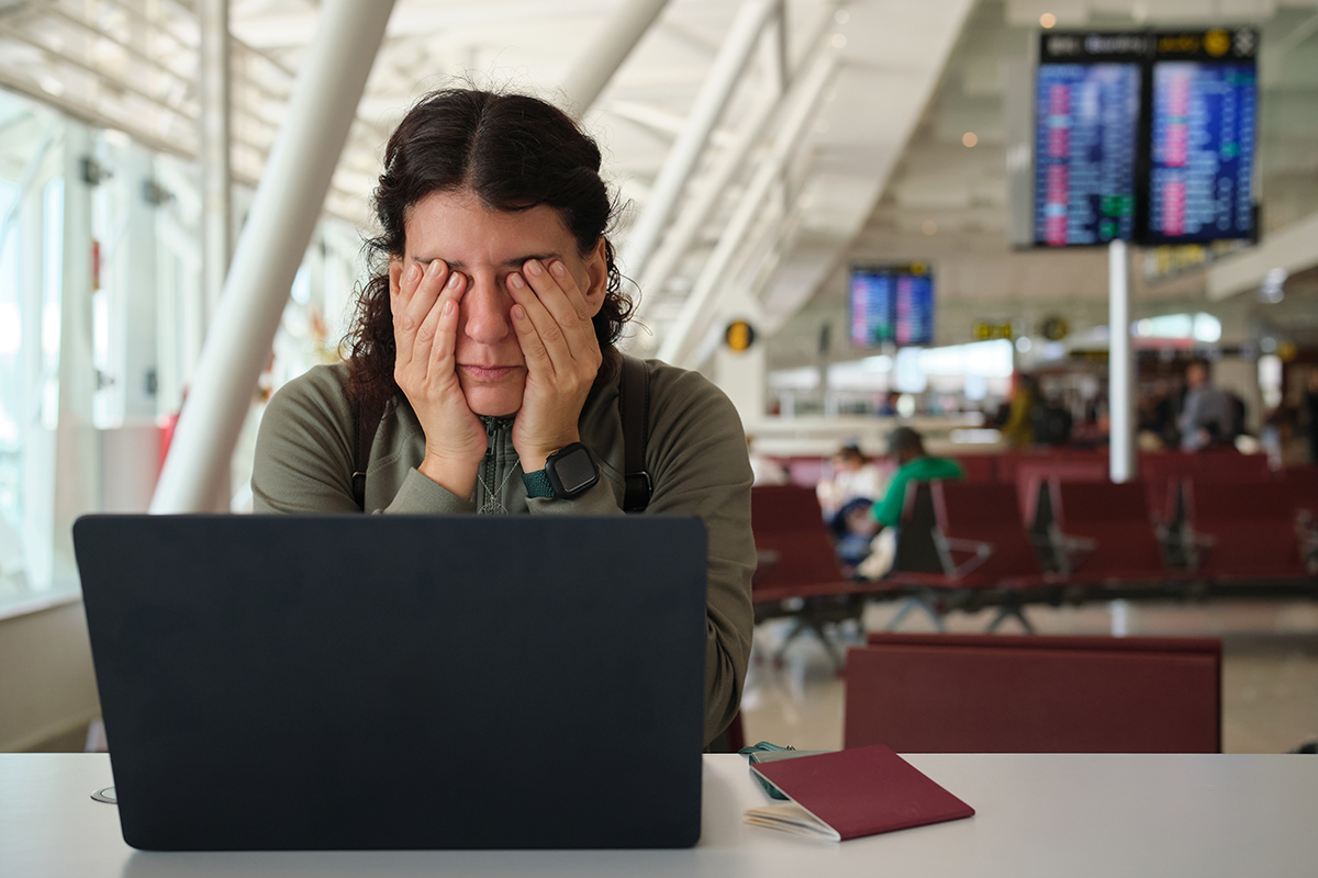 A traveler sits at an airport with a laptop and passport, covering her eyes with both hands near flight information screens.